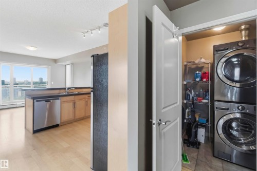 Dedicated laundry closet with stacked appliances and shelving, complemented by an open-concept kitchen featuring wood-finish cabinetry, stainless steel dishwasher, and a breakfast bar - 901 10504 99 Avenue, Edmonton, AB 