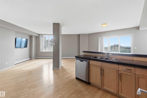 Open-concept living area featuring light wood-finish flooring, a kitchen peninsula with a double basin sink, stainless steel dishwasher, and natural wood cabinetry - 901 10504 99 Avenue, Edmonton, AB 