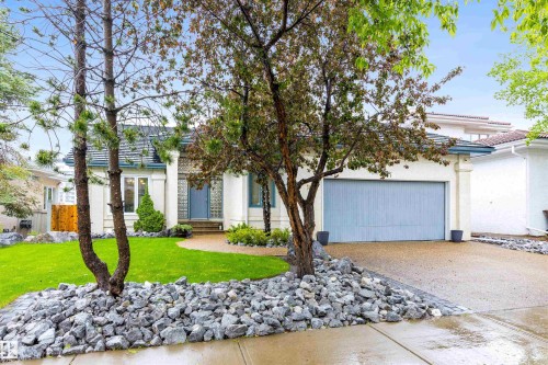 Exterior featuring a light-colored stucco finish, gray shingled roof, an attached two-car garage with a light blue door, a concrete driveway, and a front yard with manicured lawn and decorative rock landscaping - 10 Everest Crescent, St. Albert, AB 