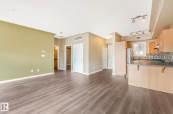 Open-concept living space featuring wood-finish flooring, a kitchen with light wood cabinetry, stainless steel refrigerator, and dark countertops - 