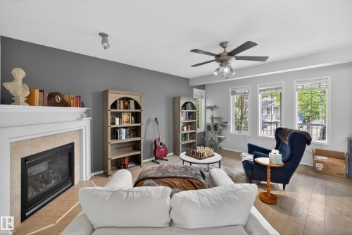 Living area featuring a fireplace with a light-toned tile surround and white mantel, wood-finish flooring, a ceiling fan with integrated lighting, and multiple windows providing natural illumination - 8 Vega Avenue, Spruce Grove, AB - Indoor Photo Showing Living Room With Fireplace