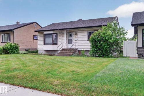 Front exterior featuring white siding, a dark asphalt shingle roof, a bay window with dark trim, an inviting front entry with railings, and a spacious green lawn - 9740 65 Avenue Nw, Edmonton, AB 