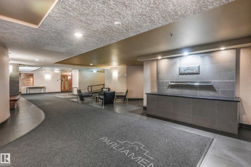 Building lobby featuring a reception desk with a dark countertop and tiled facade, contemporary wall sconces, recessed ceiling lighting, and a combination of carpet and hard-surface flooring - 1407 9939 109 Street, Edmonton, AB 