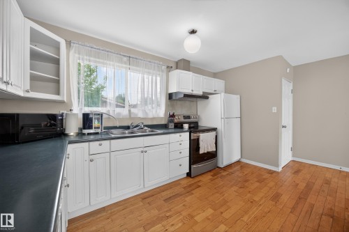 Kitchen featuring wood-finish flooring, white cabinetry, dark countertops, a double basin sink, and a large window - 5216 104A Street, Edmonton, AB - Indoor Photo Showing Kitchen With Double Sink