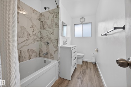 Bathroom featuring a white panel-sided bathtub with a tiled surround, wood-finish flooring, a white vanity with black hardware, and a frosted glass window - 5216 104A Street, Edmonton, AB - Indoor Photo Showing Bathroom