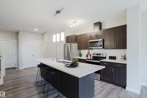 Open-concept kitchen featuring an island with pendant lighting, dark wood-finish cabinetry, stainless steel appliances, white subway tile backsplash, and light wood-finish flooring - 9142 Pear Drive, Edmonton, AB - Indoor Photo Showing Kitchen With Upgraded Kitchen