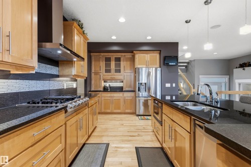 Spacious kitchen featuring natural wood cabinetry, dark countertops, a stainless steel gas cooktop with coordinating range hood, and a large central island with an undermount sink - 5709 Mcluhan Bay, Edmonton, AB 