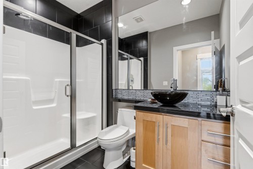 Bathroom featuring a glass-enclosed shower with integrated seating, a black vessel sink, a black countertop, a mosaic tile backsplash, and light wood cabinetry with brushed nickel hardware - 5709 Mcluhan Bay, Edmonton, AB 
