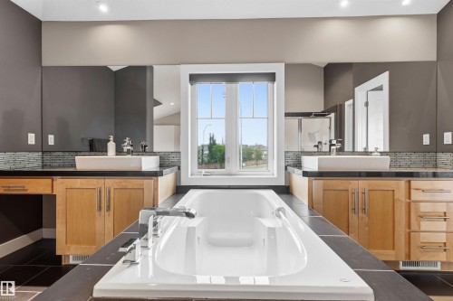 Soaking tub with integrated fixtures, dual vessel sinks, wood cabinetry, dark tile flooring, and a large window - 5709 Mcluhan Bay, Edmonton, AB 