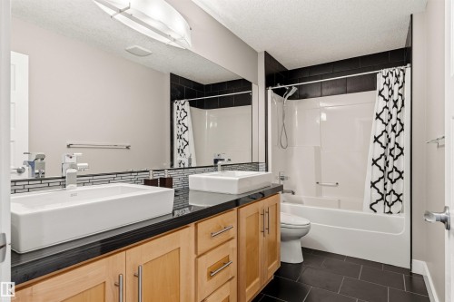 Dual vessel sinks set on a black countertop, natural wood-finish cabinetry, a large mirror, and a full bathtub with a shower featuring black tile surround - 5709 Mcluhan Bay, Edmonton, AB 