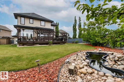 Expansive backyard featuring a large wooden deck with pergola, manicured lawn, and a decorative pond surrounded by river rock and red mulch - 5709 Mcluhan Bay, Edmonton, AB 