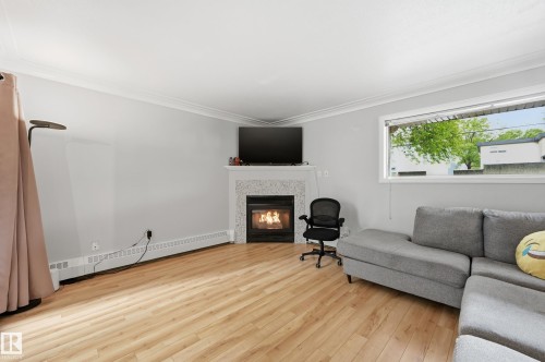 Living space featuring wood-finish flooring, a white trim fireplace with a tiled surround, a large window providing natural light, crown molding, and light gray wall paint - 33 9914 80 Avenue, Edmonton, AB - Indoor Photo Showing Living Room With Fireplace