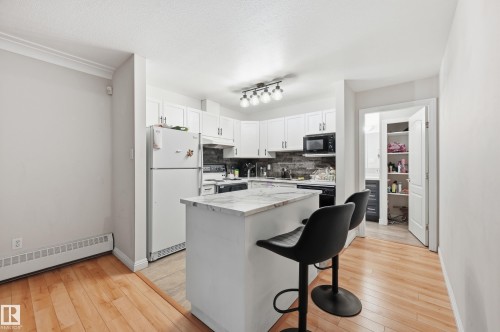 Kitchen featuring light wood-finish flooring, white cabinetry with dark hardware, a stone-look backsplash, track lighting, and a kitchen island with a stone-look countertop - 33 9914 80 Avenue, Edmonton, AB - Indoor Photo Showing Kitchen