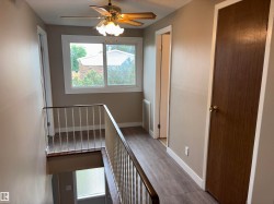 Upper-level landing featuring wood-finish flooring, a ceiling fan with integrated lighting, a large window, white trim, and a warm neutral wall color - 