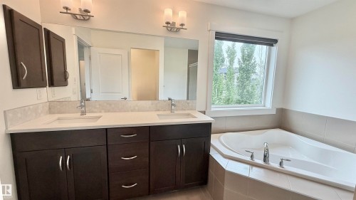 Bathroom featuring a dual vanity with dark wood cabinetry and light countertops, a large framed mirror, and an integrated corner bathtub with tiled surround - 3962 Claxton Loop, Edmonton, AB - Indoor Photo Showing Bathroom