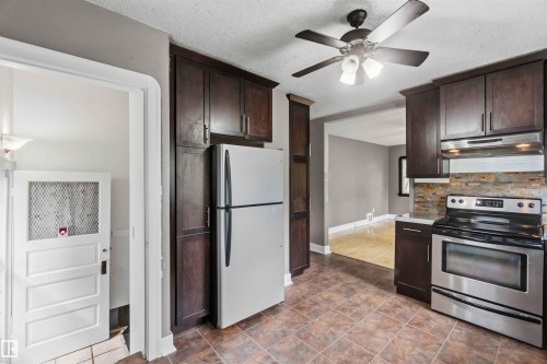 Kitchen featuring square tile flooring, dark wood-finish cabinetry, stainless steel appliances, a multi-tone stone backsplash, and a ceiling fan with integrated lighting - 11842 93 Street, Edmonton, AB 