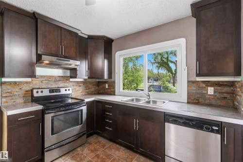 Kitchen featuring dark wood-finish cabinetry with modern hardware, stainless steel appliances, a stacked stone backsplash, and light-toned countertops - 11842 93 Street, Edmonton, AB 