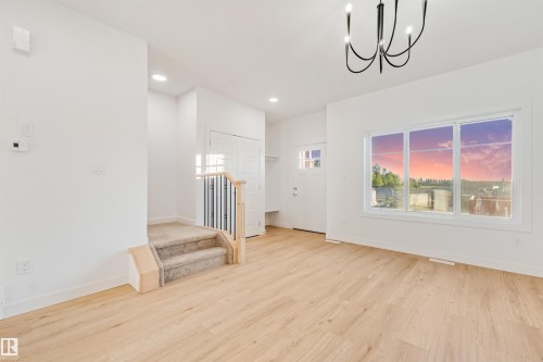 Light wood-finish flooring, white walls, modern chandelier, large multi-pane window, and carpeted stairs with a metal and wood railing - 1511 211 Street Nw, Edmonton, AB 