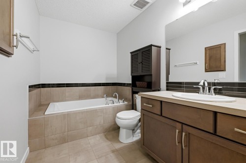 Bathroom featuring a built-in soaking tub, tiled surround, wood-finish vanity with countertop sink, large vanity mirror, and dark wood-finish storage cabinet - 17844 61 Street, Edmonton, AB 