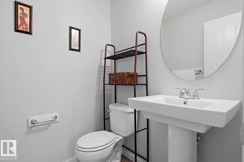Pedestal sink with chrome faucet, circular mirror, and adjacent toilet with an open-shelf storage unit - 17844 61 Street, Edmonton, AB 