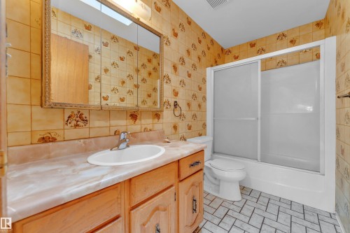 Bathroom featuring a wood vanity with an integrated sink, a large mirrored medicine cabinet, a full bathtub with sliding glass doors, and a tiled floor - 10819 54 Avenue, Edmonton, AB 