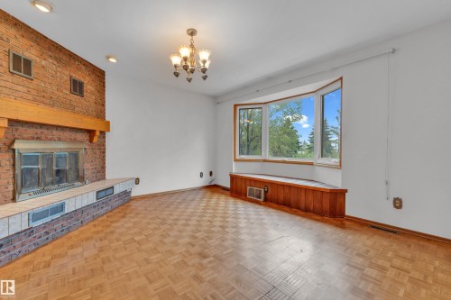 Spacious room featuring a brick fireplace with a wood mantle, an expansive bay window with a wood-paneled window seat, parquet wood-finish flooring, and a decorative chandelier - 10819 54 Avenue, Edmonton, AB 