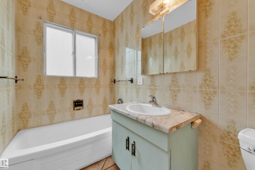 Bathroom featuring patterned wall tiles, a white bathtub, a window with obscure glass, a vanity with a counter-mounted sink, and a mirrored medicine cabinet - 10819 54 Avenue, Edmonton, AB 