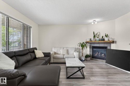 Spacious living area featuring wood-finish flooring, a corner fireplace with stone surround and wooden mantel, and a large window with horizontal blinds - 21244 90 Avenue, Edmonton, AB 