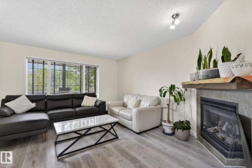 Living area featuring wood-finish flooring, a corner fireplace with tile surround, a wood mantel, a large window with blinds, and a ceiling-mounted light fixture - 21244 90 Avenue, Edmonton, AB 
