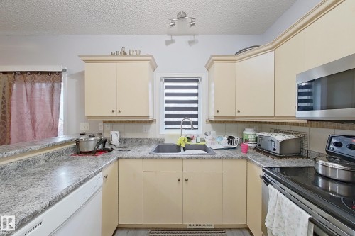 Kitchen featuring cream cabinetry, stainless steel microwave, integrated dishwasher, dark composite sink, and grey flecked countertops - 41 5102 30 Avenue, Beaumont, AB 