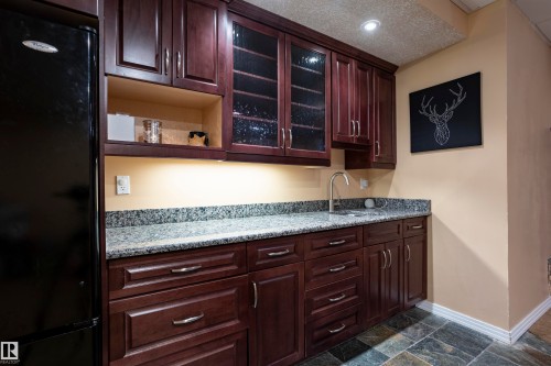 Kitchenette featuring dark wood-finish cabinetry with glass-front uppers, granite countertops, an undermount sink with a gooseneck faucet, and slate tile flooring - 1441 Highwood Boulevard, Devon, AB 