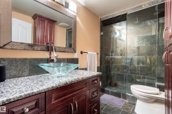 Bathroom featuring a glass vessel sink, granite countertop, rich wood-finish vanity, framed mirror, and a glass-enclosed shower with stone-look tile - 