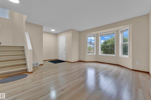 Bright living area featuring light wood-finish flooring, a bay window with multiple panes, neutral wall tones, and a staircase with white risers and handrail - 3134 30 Avenue Nw, Edmonton, AB 