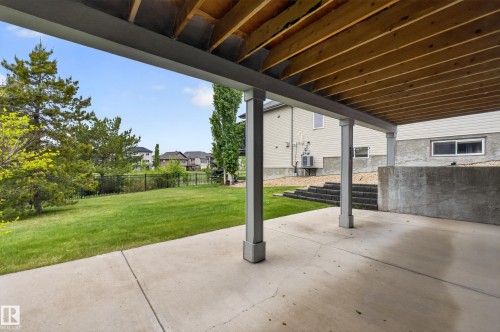 Covered concrete patio with wood-finish ceiling, featuring a manicured lawn, partial fencing, and concrete retaining walls - 624 Cantor Landing Landing, Edmonton, AB 