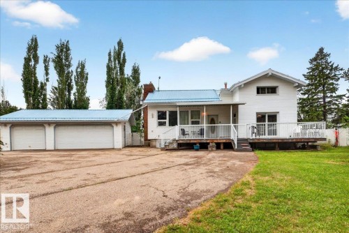 White exterior with a light blue metal roof, featuring a multi-car garage, expansive driveway, and a large deck with white railings - 140 50047 Rge Rd 244, Rural Leduc County, AB 