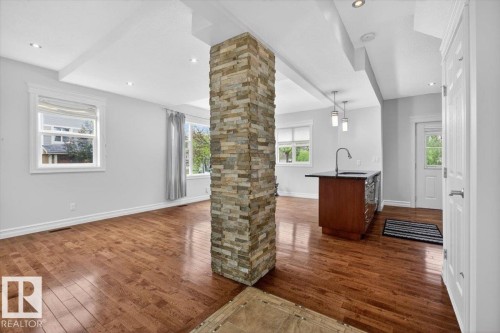 Open-concept interior featuring wood-finish flooring, a stacked stone accent column, recessed lighting, white trim and baseboards, and a kitchen island with a dark countertop and pendant lighting - 10958 88 Avenue, Edmonton, AB 