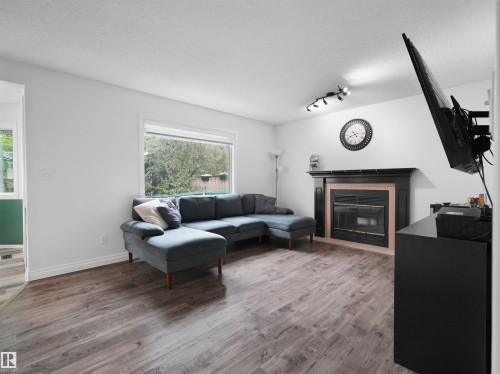 Inviting living area featuring wood-finish flooring, a prominent fireplace with a black mantel, a large window, and track lighting - 208 Ferguson Place, Edmonton, AB 