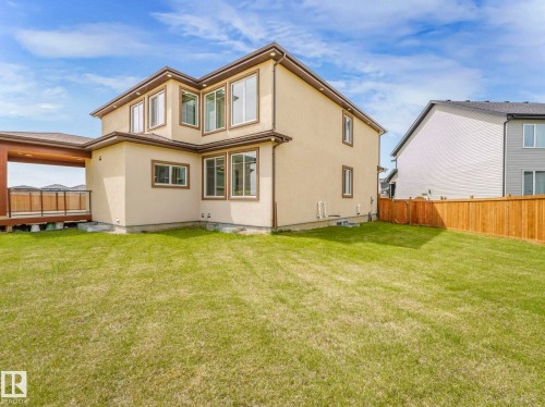 Expansive rear exterior featuring stucco siding, multiple large windows, a covered deck with dark railing, a wooden privacy fence, and a spacious lawn area - 3101 Pelerin Crescent, Beaumont, AB 