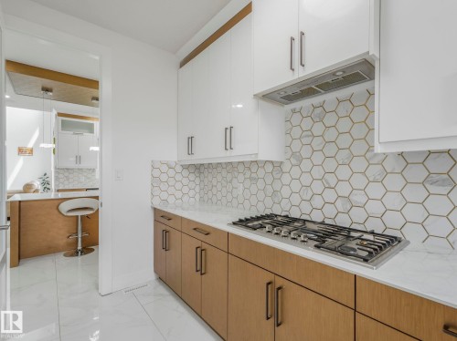 Modern kitchen featuring a gas cooktop, sleek range hood, white shaker upper cabinetry, wood-finish lower cabinetry, and a geometric tile backsplash with gold-tone accents - 3101 Pelerin Crescent, Beaumont, AB 