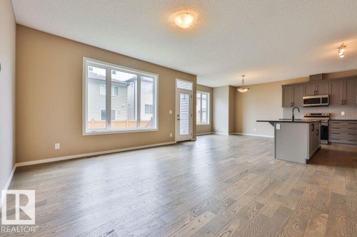 Open-concept living space featuring wood-finish flooring, large windows, a kitchen island with seating, dark shaker-style cabinetry, and stainless steel appliances - 2031 207 Street, Edmonton, AB 