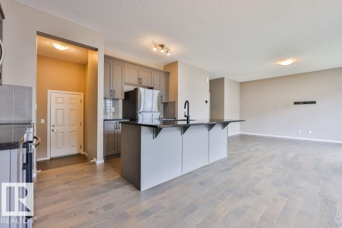 Open-concept living space featuring wood-finish flooring, a kitchen island with seating, a flat-panel cabinetry, a stainless steel refrigerator, and a tile backsplash - 2031 207 Street, Edmonton, AB 