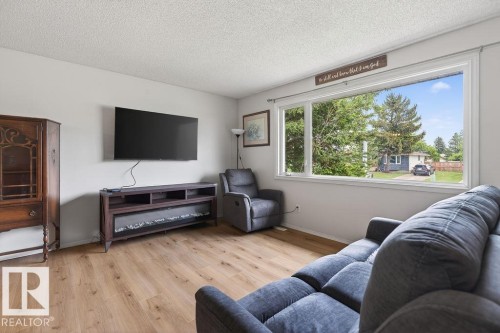 Living room featuring expansive windows, wood-finish flooring, a wall-mounted entertainment unit with an electric fireplace, light neutral wall paint, and a textured white ceiling - 123 Grandin Drive, Morinville, AB 