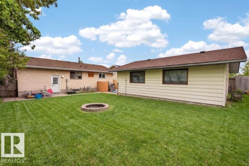 Expansive green lawn with a circular stone fire pit, bordered by a stucco-finished exterior featuring a white entry door and multiple windows - 123 Grandin Drive, Morinville, AB 