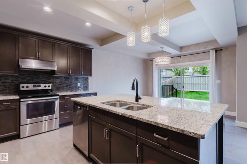 Kitchen featuring an island with a dual-basin sink and a matte black faucet, dark wood-finish cabinetry, stainless steel appliances, and light-toned granite countertops - 1957 Ainslie Link, Edmonton, AB 