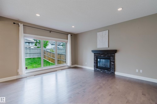 Spacious room featuring wood-finish flooring, a contemporary stacked stone fireplace with a dark mantel, and recessed ceiling lighting - 1957 Ainslie Link, Edmonton, AB 