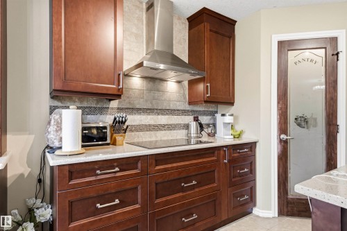 Kitchen featuring rich wood cabinetry, light-toned countertops, a stainless steel range hood, an integrated cooktop, and a glass-paneled pantry door - 32 Lamplight Bay, Spruce Grove, AB 