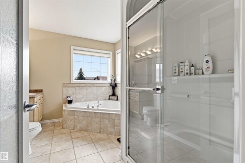 Bathroom featuring a spacious corner soaking tub with tiled surround, a separate glass-enclosed shower, a double vanity with stone-finish countertops, and tiled flooring - 32 Lamplight Bay, Spruce Grove, AB 