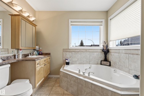 Spacious bathroom featuring a corner soaking tub with tiled surround, a light wood-finish vanity with an integrated sink and ample counter space, multiple windows, and ceramic tile flooring - 32 Lamplight Bay, Spruce Grove, AB 