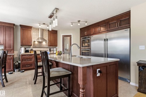 Spacious kitchen featuring an expansive center island with an integrated sink, rich wood cabinetry, stainless steel appliances, a range hood, and tile flooring - 32 Lamplight Bay, Spruce Grove, AB 