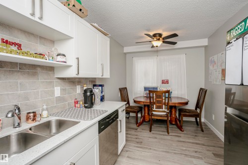 Kitchen and dining area featuring white cabinetry with silver hardware, light grey subway tile backsplash, a double basin stainless steel sink, wood-finish flooring, and a ceiling fan with integrated lighting - 3103 109 Street, Edmonton, AB 
